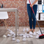 Low Section Of Female Janitor Sweeping Crumbled Paper On Floor With Broom