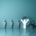 A line of stick men against a wall. One is illuminated with its arms raised as a magnifying glass hovers over it