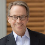 Headshot of Hamilton Lane wearing black blazer, light shirt and glasses against paneled background