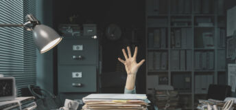 Image of a person's hand reaching out from under a desk covered in a pile of paper work and empty mugs.
