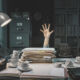 Image of a person's hand reaching out from under a desk covered in a pile of paper work and empty mugs.